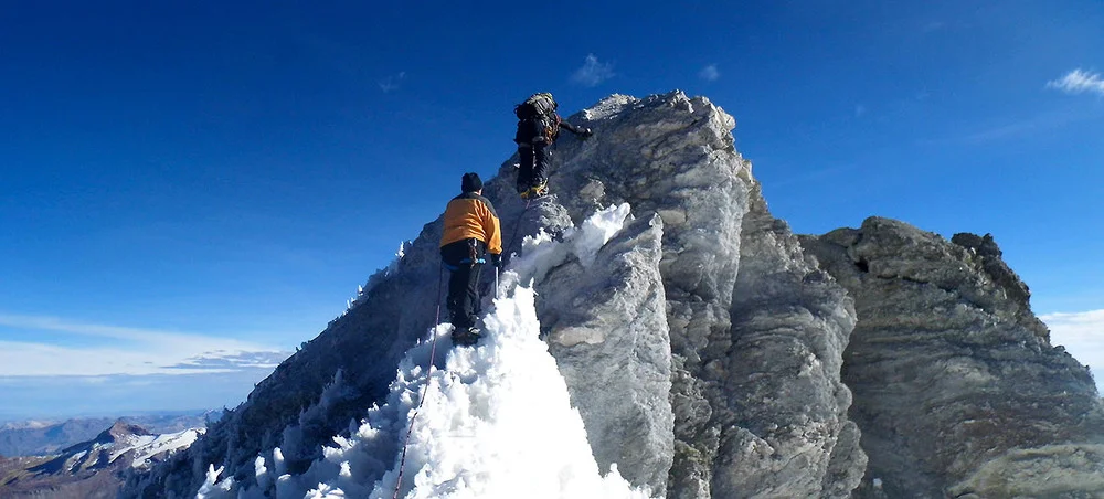 Two hikers standing on the snowy summit of Ampato Volcano, surrounded by bright white glaciers and expansive Andean views. | Andean Travel Experience