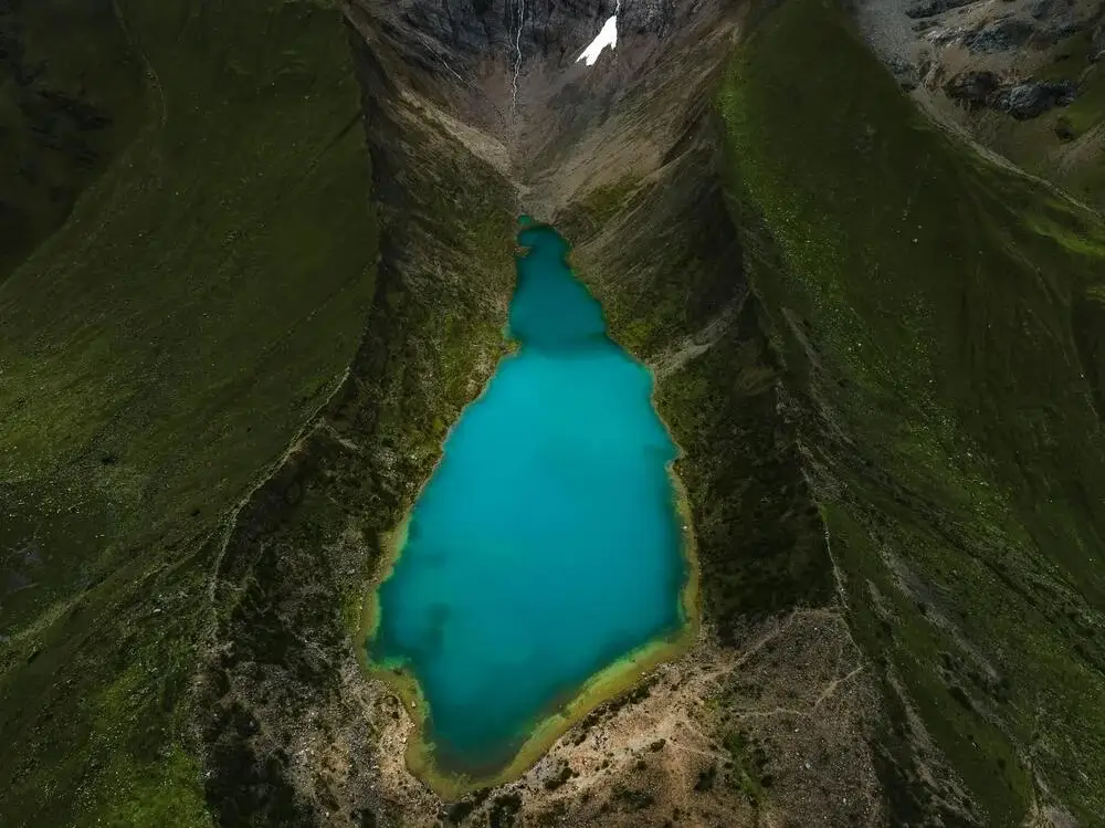 Turquoise Humantay Lake surrounded by towering snow-capped mountains and rocky Andean terrain on a bright, clear day. | Andean Travel Experience