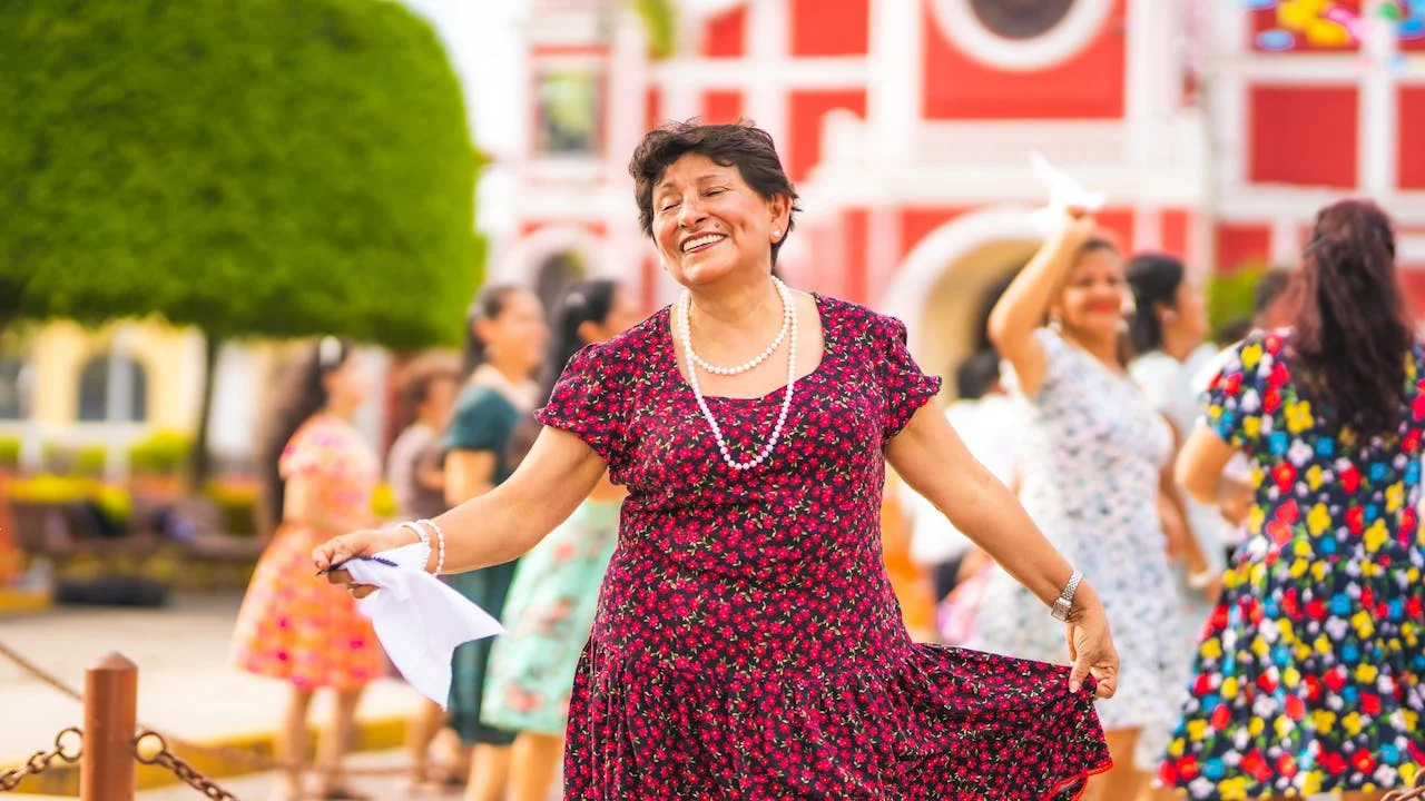 A joyful Peruvian woman dances in a traditional festival, surrounded by other women in floral dresses, each holding a handkerchief — a typical element in many folkloric Peruvian dances. Her bright smile and vibrant attire reflect the spirit of Peruvian culture: colorful, festive, and full of life. In the background, trimmed trees and a colonial-style church set the scene in a lively town square, capturing the essence of Peru's cultural heritage. | Andean Travel Experience