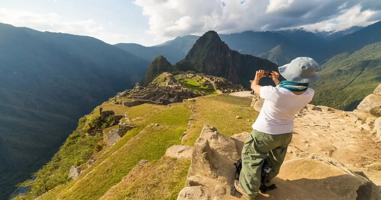 A tourist taking photos of Machu Picchu, Peru, with the ancient Inca ruins and surrounding mountains in the background | Andean Travel Experience